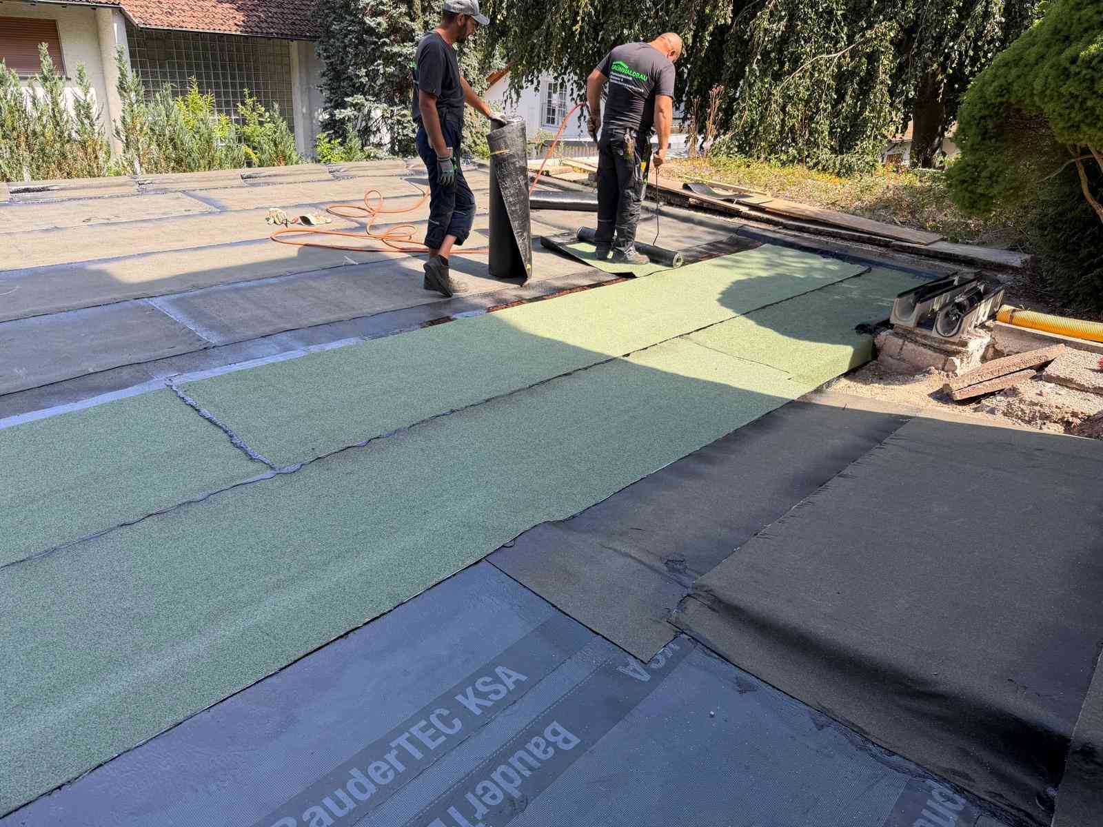 Zwei Mitarbeiter von Grünwaldbau verlegen und verschweißen Bitumenbahnen auf einer Flachdachfläche im Sonnenlicht